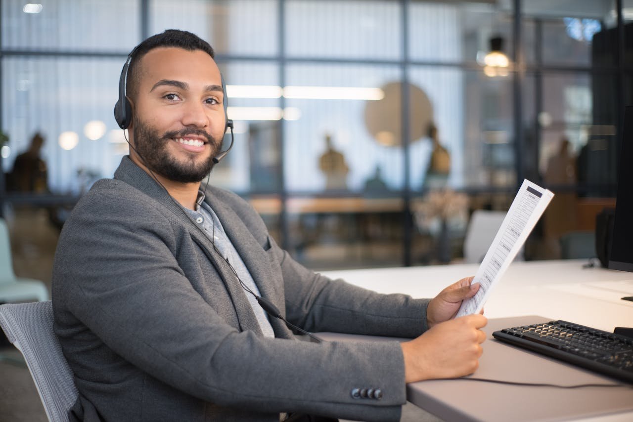 Smiling man wearing a headset, holding a document, sitting at an office desk.
